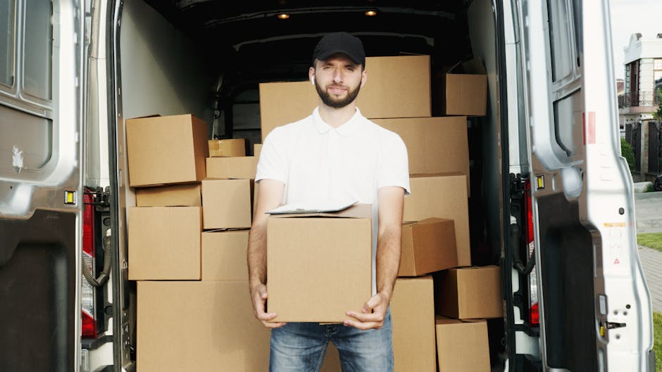 A man with a beard, dressed in a white polo shirt, jeans, and a black cap, standing next to the open rear doors of a moving van in Wembley. Inside the van, there are numerous cardboard boxes of varyin