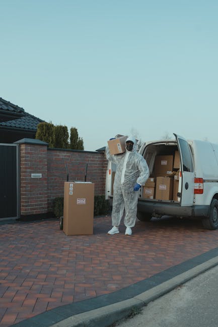 A mover dressed in a white protective suit, gloves, and a face mask is seen standing on a brick driveway next to a partially open white van used for home relocation within the Wembley area. The mover
