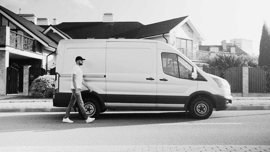 A mover dressed in a white protective suit, gloves, and a face mask is seen standing on a brick driveway next to a partially open white van used for home relocation within the Wembley area. The mover