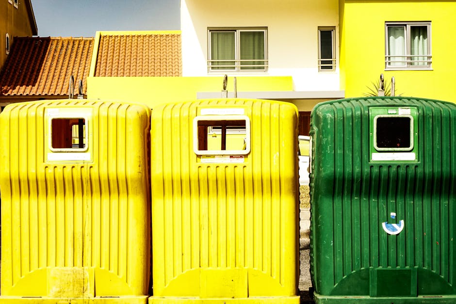 A group of purple and white portable toilet units arranged outdoors on a grassy area with small yellow flowers. The units are positioned in multiple rows, with some on the grass and others limited to 
