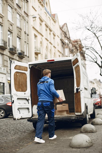A man wearing a blue jacket, dark trousers, and white footwear is seen loading cardboard boxes into the back of a white panel van parked on a cobbled street in a residential area. The van, which is as