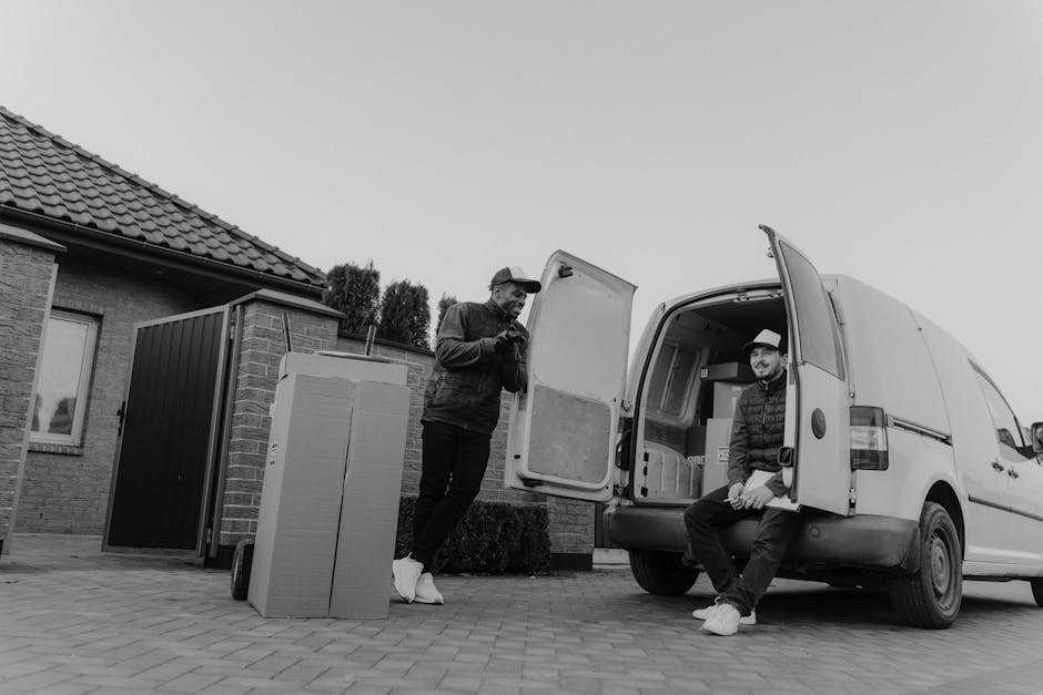 A man wearing a blue jacket, dark trousers, and white footwear is seen loading cardboard boxes into the back of a white panel van parked on a cobbled street in a residential area. The van, which is as