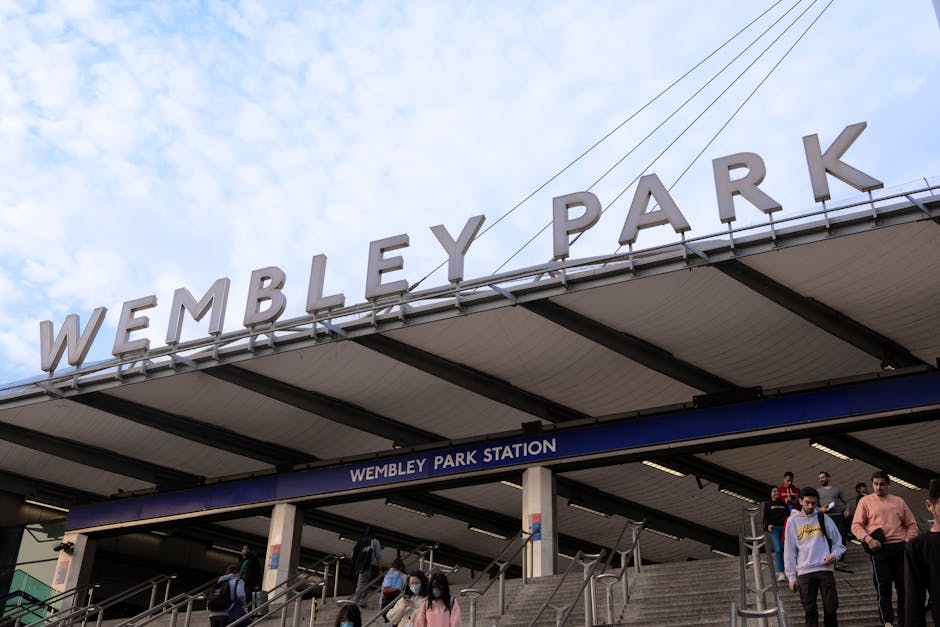 Outside view of Wembley Park Station with a large metal sign spelling 'WEMBLEY PARK' mounted on the roof structure. Below, there are multiple people walking on the wide stone staircase leading into the station entrance, some wearing face masks and carrying bags. The station's blue signage with white text 'WEMBLEY PARK STATION' is mounted above the staircase. The station's exterior has a modern, functional design with a metal roof supported by black beams. In the background, the sky is partly cloudy with patches of blue visible, indicating a daytime setting. This scene illustrates a busy transportation hub often associated with home relocation and moving logistics in Wembley, reflecting the importance of efficient furniture transport and packing and moving services, such as those provided by Man With a Van Wembley.