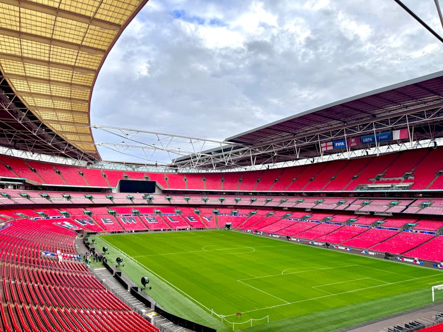 A large, modern stadium with a partially open roof, featuring rows of red seating surrounding a well-maintained grass football pitch with white goalposts at each end. Inside the stadium, there are several black equipment cases and lighting fixtures positioned along the perimeter of the pitch, indicating preparations for a match or event, as well as white ropes and barriers. The interior environment shows a mix of metal framework and fabric roofing, with natural daylight illuminating the space. The scene may reflect the site of a home relocation or moving service, such as those offered by Man With a Van Wembley, involving the transportation of equipment or furnishings during a property move, with the stadium serving as a backdrop for logistics planning or loading activities.