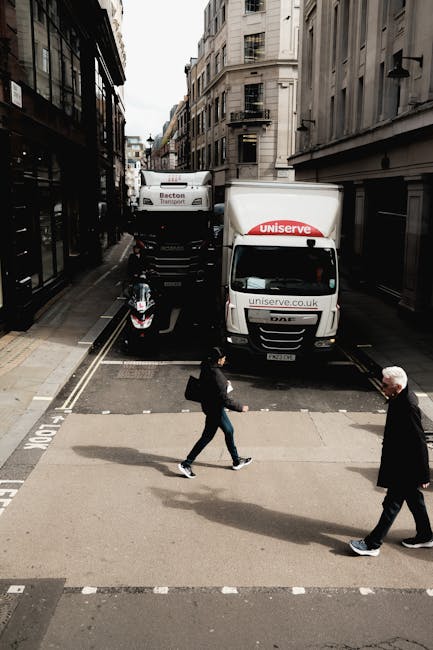A small white moving van with a closed cargo area is parked on a city street in front of an ornate, multi-storey building with a rounded corner, large windows, and decorative stonework. To the left, t